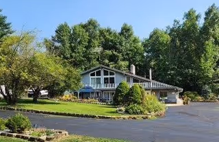 A single-story residential building with large windows and a sloped roof, surrounded by green trees and a well-maintained lawn with shrubs. The driveway is paved and curves around the front of the building.