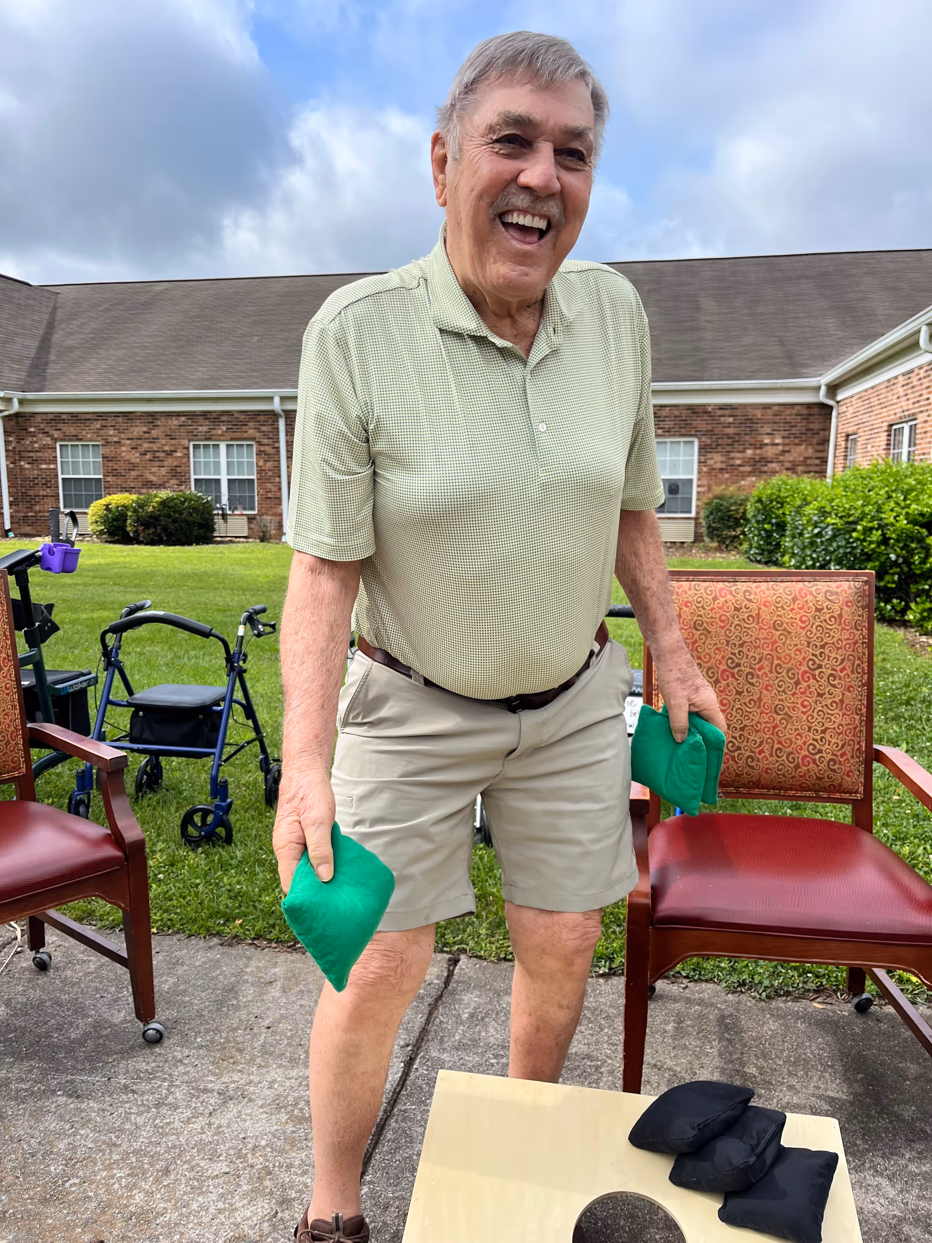 An elderly man standing outdoors on a paved area between two chairs, holding green bean bags in both hands, smiling and appearing to play a bean bag toss game. Behind him is a grassy lawn with a walker and a brick building under a partly cloudy sky.