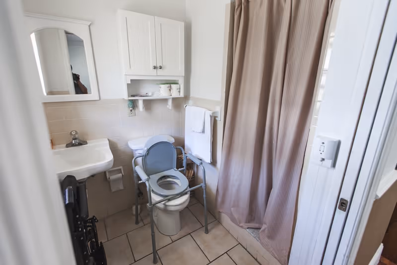 A small bathroom with a toilet equipped with a raised seat and safety frame, a white sink, a wall-mounted cabinet above the toilet, a towel rack with a white towel, and a shower with a beige curtain. The floor is tiled and the walls are partially tiled in beige.
