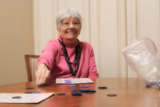An elderly woman with white hair and glasses, wearing a pink sweater and black beaded necklace, is sitting at a wooden table playing a game with blue chips and bingo cards. She is smiling and reaching out to place a chip on the table. A plastic bag is visible on the right side of the table.