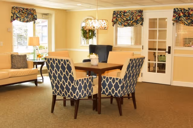 A cozy interior common area with a square wooden table surrounded by four upholstered chairs, two with a blue and white geometric pattern and two in solid beige. In the background, there is a beige sofa with a dark cushion, a side table with a lamp and flowers, a window with floral valances, a decorative mirror with a wreath, and a white door with glass panes.