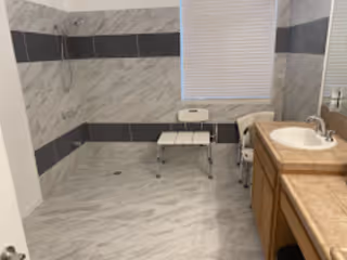 Spacious accessible bathroom with marble-look tile, roll-in shower with handheld head and bench, a single-sink vanity with countertop, and a window with blinds.