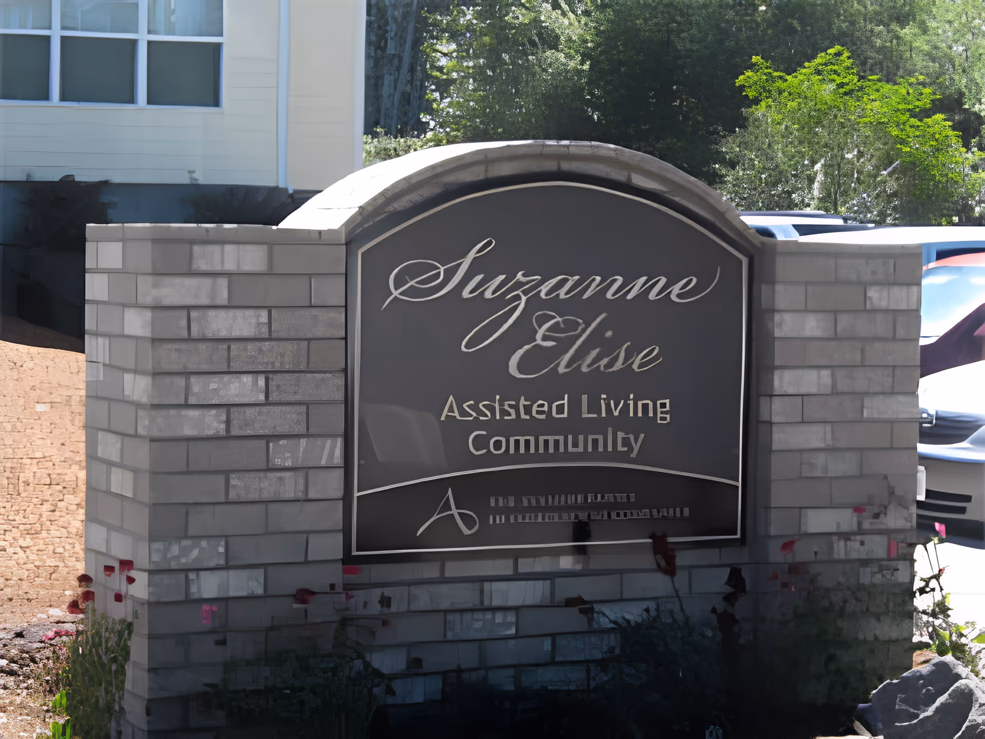 Stone entry sign that reads "Suzanne Elise Assisted Living Community" with a building, cars, and trees in the background.