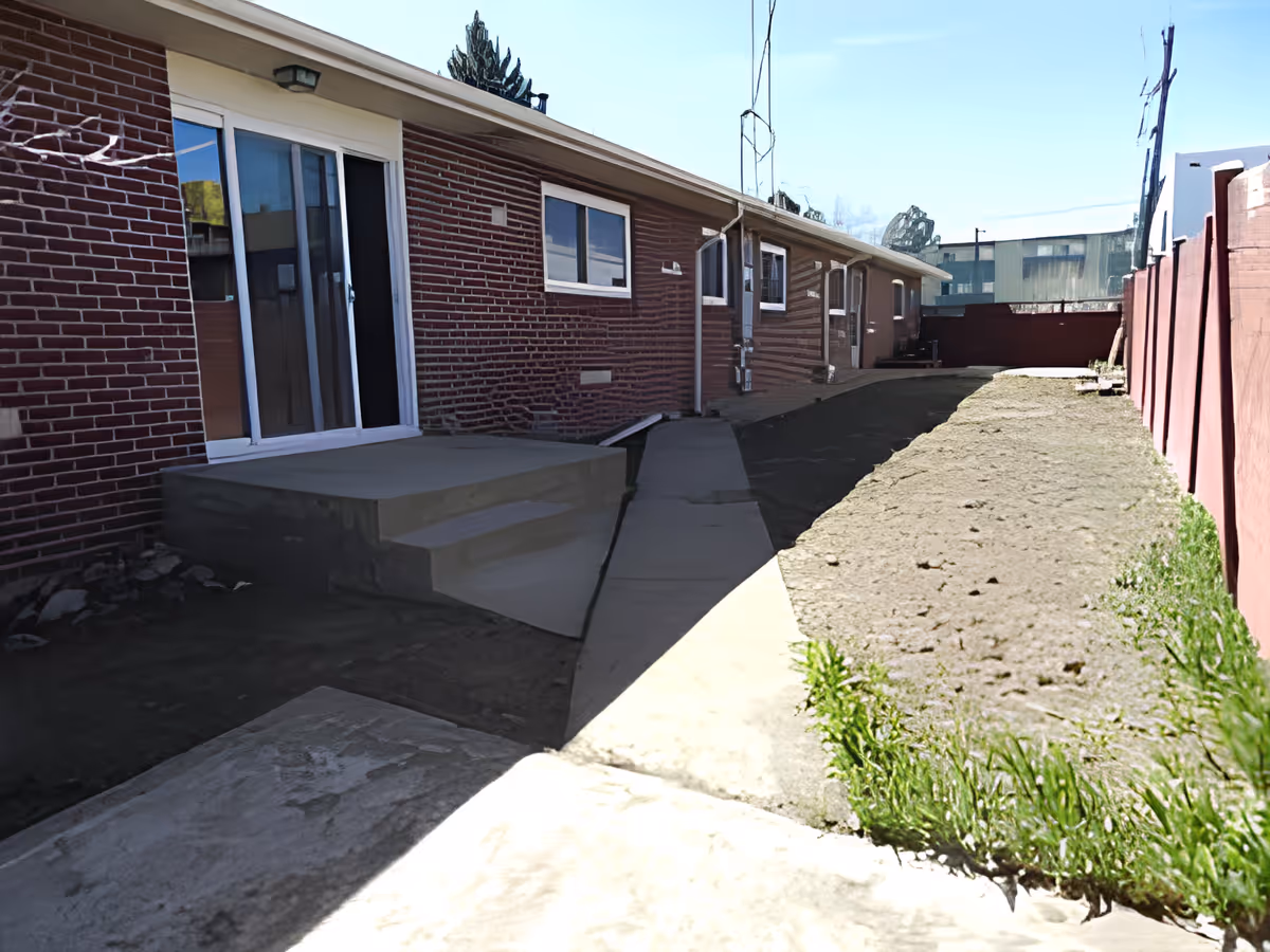 Outdoor view of a narrow backyard area beside a brick building with sliding glass doors and windows. There is a concrete pathway and steps leading to the doors, with a dirt and sparse grass area on the right side, bordered by a red fence.