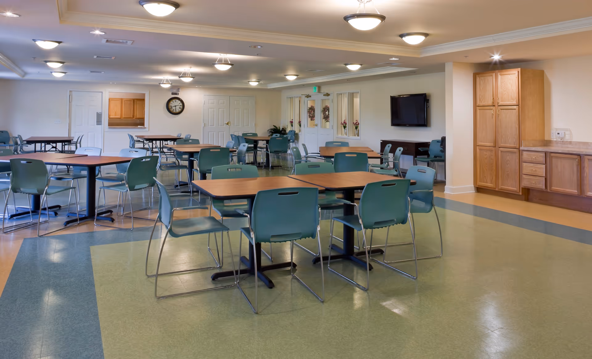 A spacious dining room with multiple wooden tables and green chairs arranged neatly. The room has a tiled floor with green and blue patterns, ceiling lights, a wall clock, a mounted TV, and wooden cabinets along one wall. Double doors and a serving window are visible in the background.