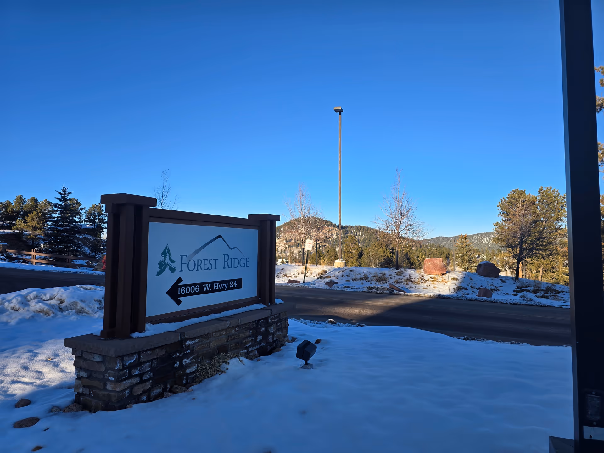 Outdoor scene showing a sign for Forest Ridge Senior Living with the address 16006 W. Hwy 24. The sign is mounted on a stone base and is surrounded by snow-covered ground. In the background, there are trees, a road, a streetlamp, and hills under a clear blue sky.