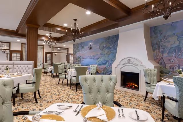 Elegant dining room with light green tufted chairs and white tablecloth-covered tables set with gold chargers, silverware, and napkins. The room features a decorative fireplace, chandeliers, wooden ceiling beams, and a large mural depicting a serene landscape with trees and a river.