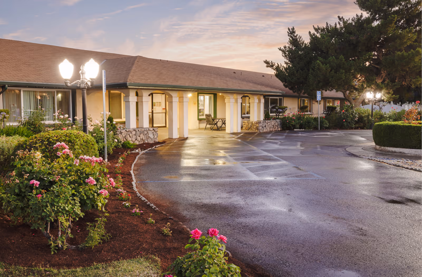 Front entrance of a single-story care facility with a curved driveway, landscaped flowerbeds, and lit lampposts at dusk.