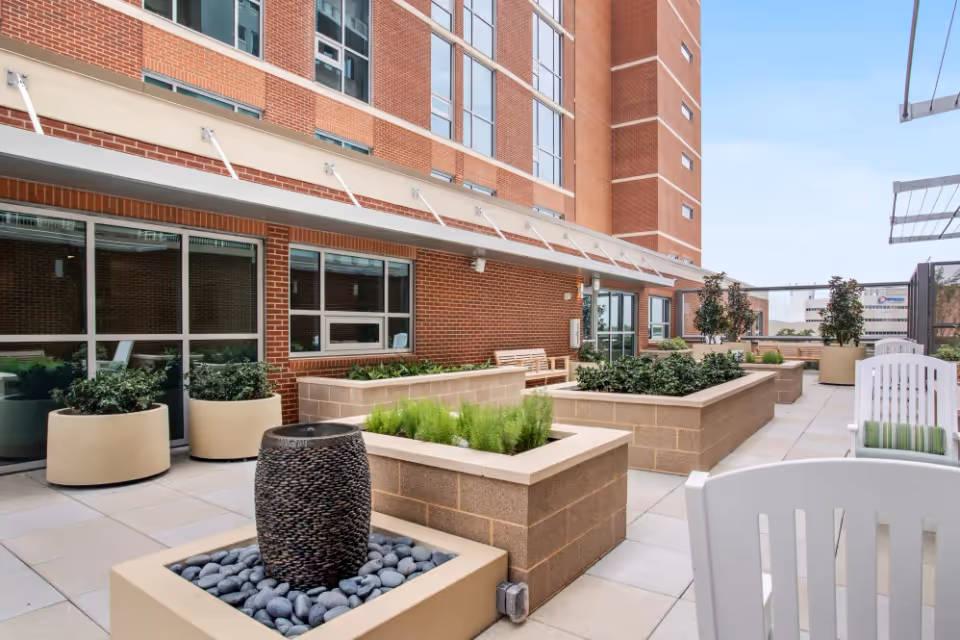 Outdoor patio area at The Village of Southampton featuring large rectangular planters with green plants, a decorative stone water feature, white chairs with striped cushions, and a brick building facade with large windows in the background under a clear blue sky.