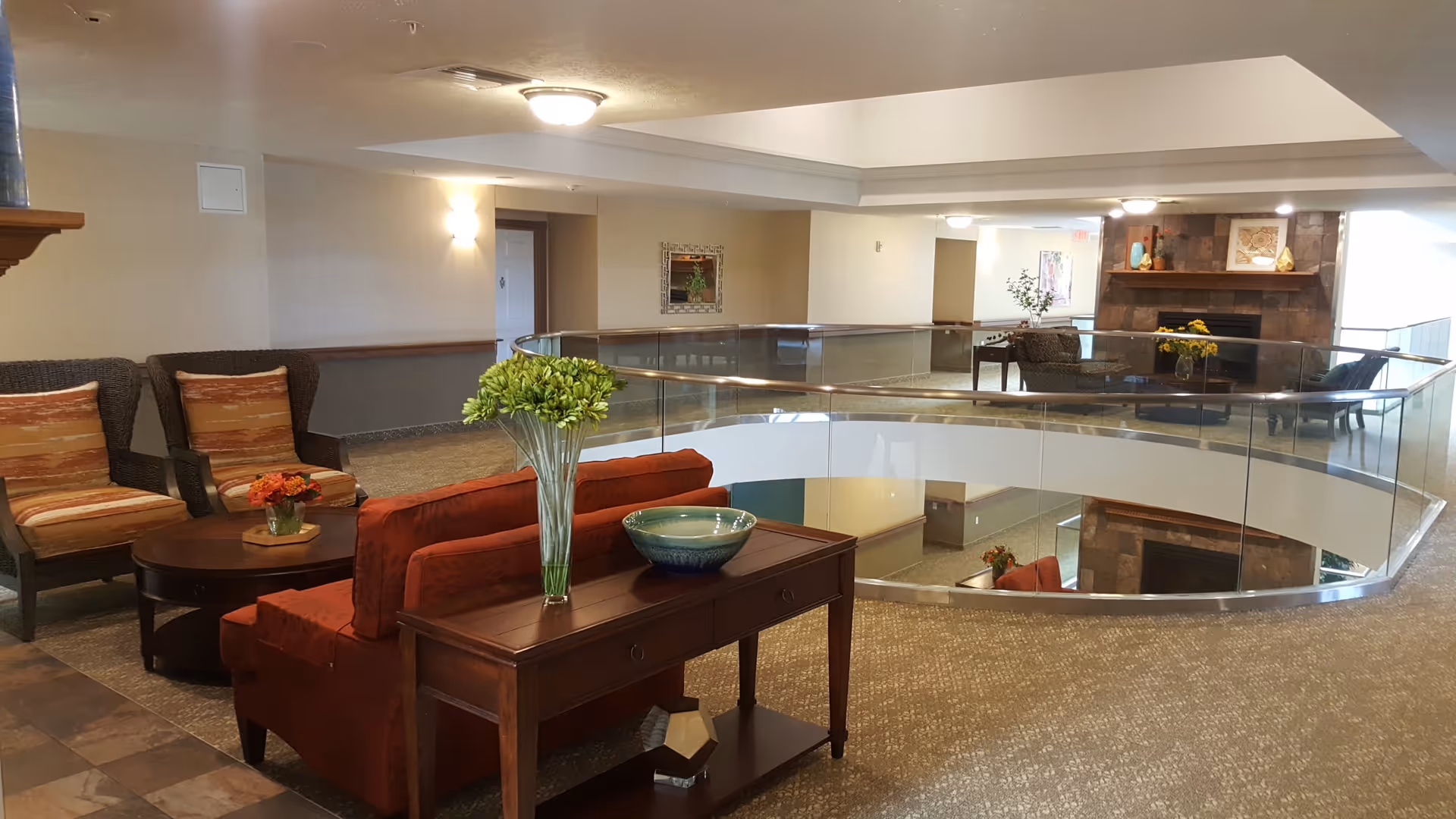 A spacious and well-lit common area in a retirement community featuring comfortable seating including a red sofa and two armchairs with striped cushions. A wooden console table with a vase of green flowers and a decorative bowl is placed behind the sofa. The area has a glass railing overlooking a lower level, and a stone fireplace with decorations is visible in the background.