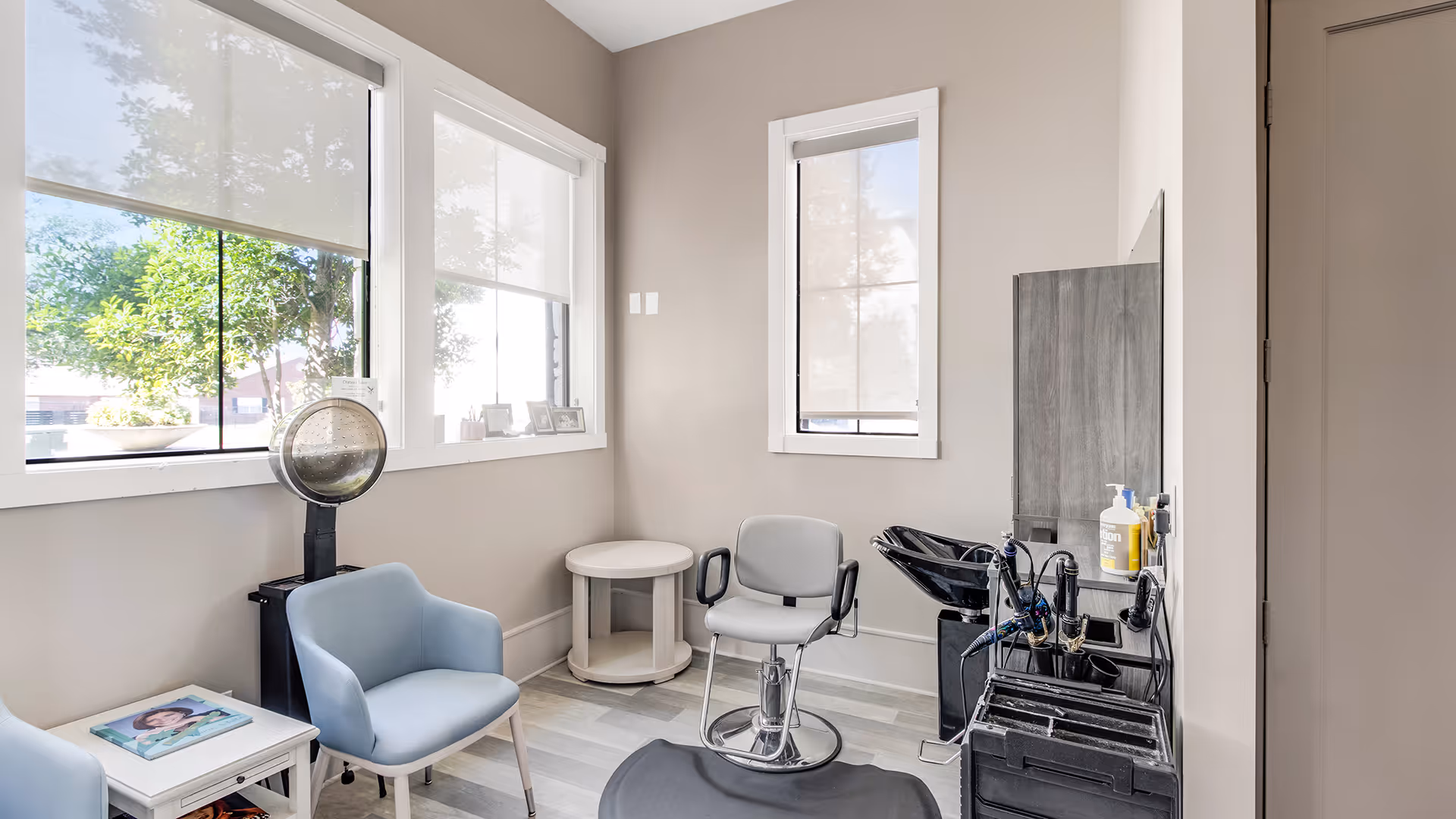 A bright and clean hair salon room with two light blue armchairs, a white side table with a magazine, a hair dryer hood, a salon styling chair in front of a black wash basin, and a wall-mounted mirror. The room has large windows with white roller blinds and light-colored walls and flooring.