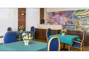 Interior view of a dining area with tables covered in green tablecloths and blue cushioned chairs. Each table has a small flower arrangement. A colorful mosaic artwork is displayed on the wall in the background.
