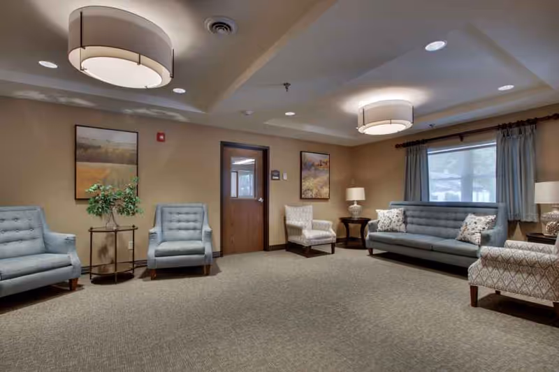 A cozy living room area in a senior care facility featuring a blue sofa with patterned pillows, two matching blue armchairs, two patterned armchairs, two side tables with lamps, a small round table with a plant, beige walls with framed artwork, a window with curtains, and ceiling lights.