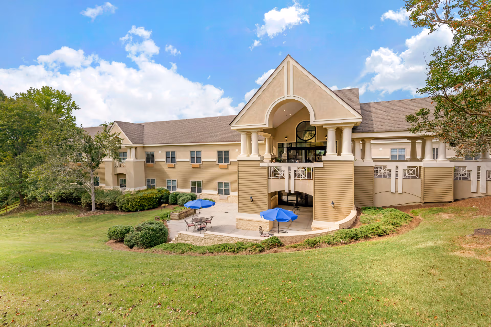 Exterior view of a large senior living facility building with beige siding and a peaked roof. The building features a covered patio area with blue umbrellas and outdoor seating. The surrounding area includes green grass, bushes, and trees under a partly cloudy blue sky.