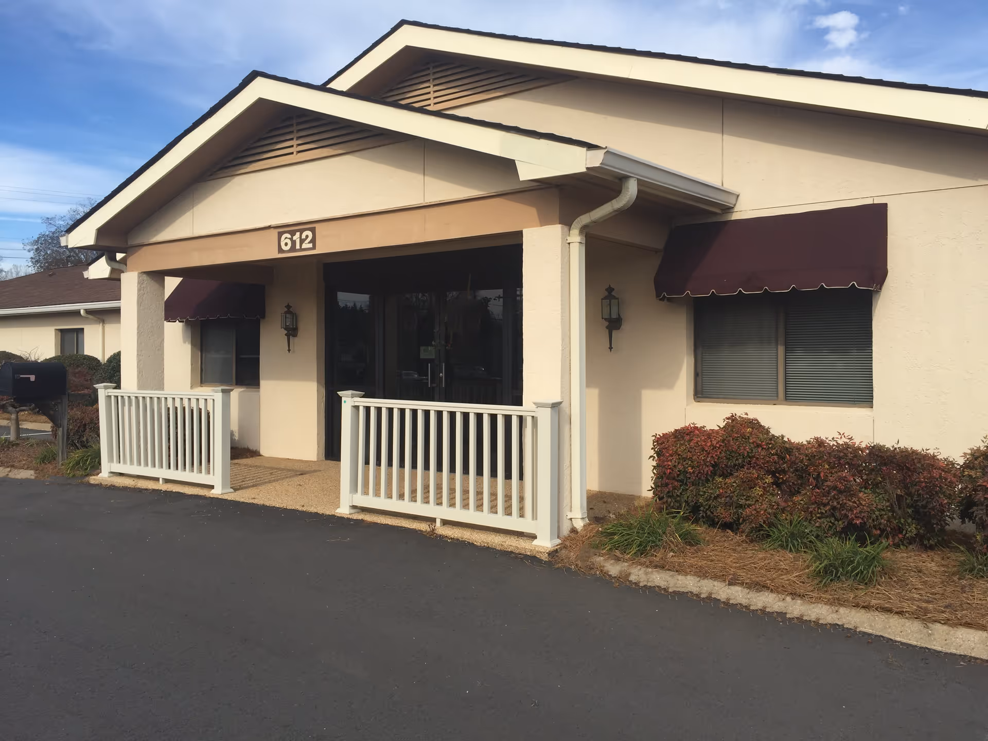 Exterior view of a single-story building with beige walls and a brown roof. The entrance has a small covered porch with white railings and the number 612 displayed above the door. There are two wall-mounted lantern-style lights on either side of the entrance and maroon awnings over the windows. Shrubs and plants are visible along the side of the building.