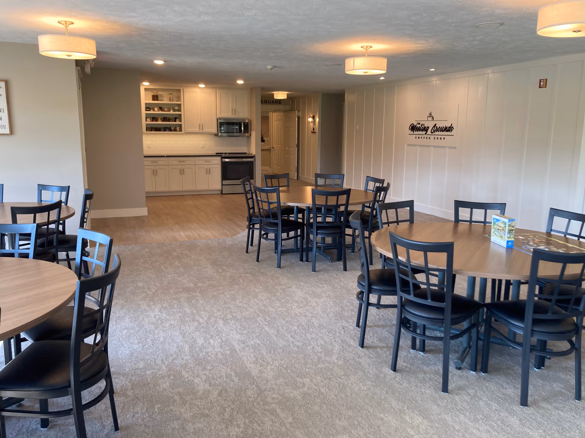 Communal dining room with multiple round tables and chairs, a kitchenette in the back, and a coffee-shop sign on the wall.