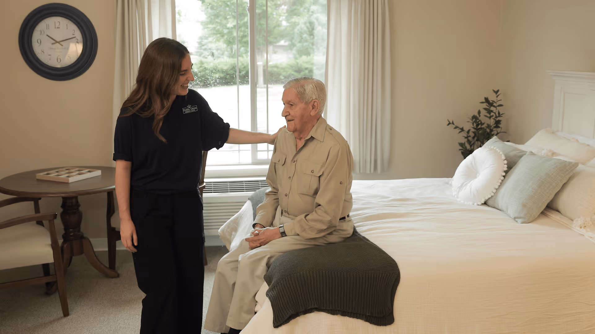 A caregiver smiling and gently touching the shoulder of an elderly man who is sitting on the edge of a neatly made bed in a well-lit bedroom with a round table and chairs, a clock on the wall, and a window with curtains.