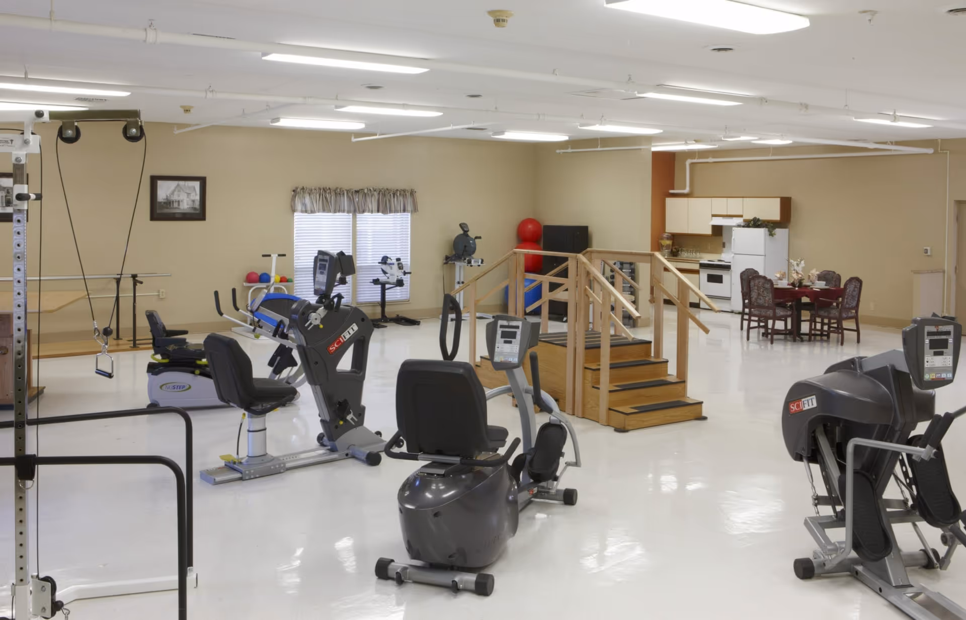 A spacious rehabilitation or exercise room with various fitness equipment including recumbent exercise bikes, resistance machines, and balance balls. There is a small wooden staircase with handrails in the center, and a kitchenette with a dining table and chairs in the background. The room has beige walls, white ceiling with fluorescent lights, and a window with blinds and a valance.