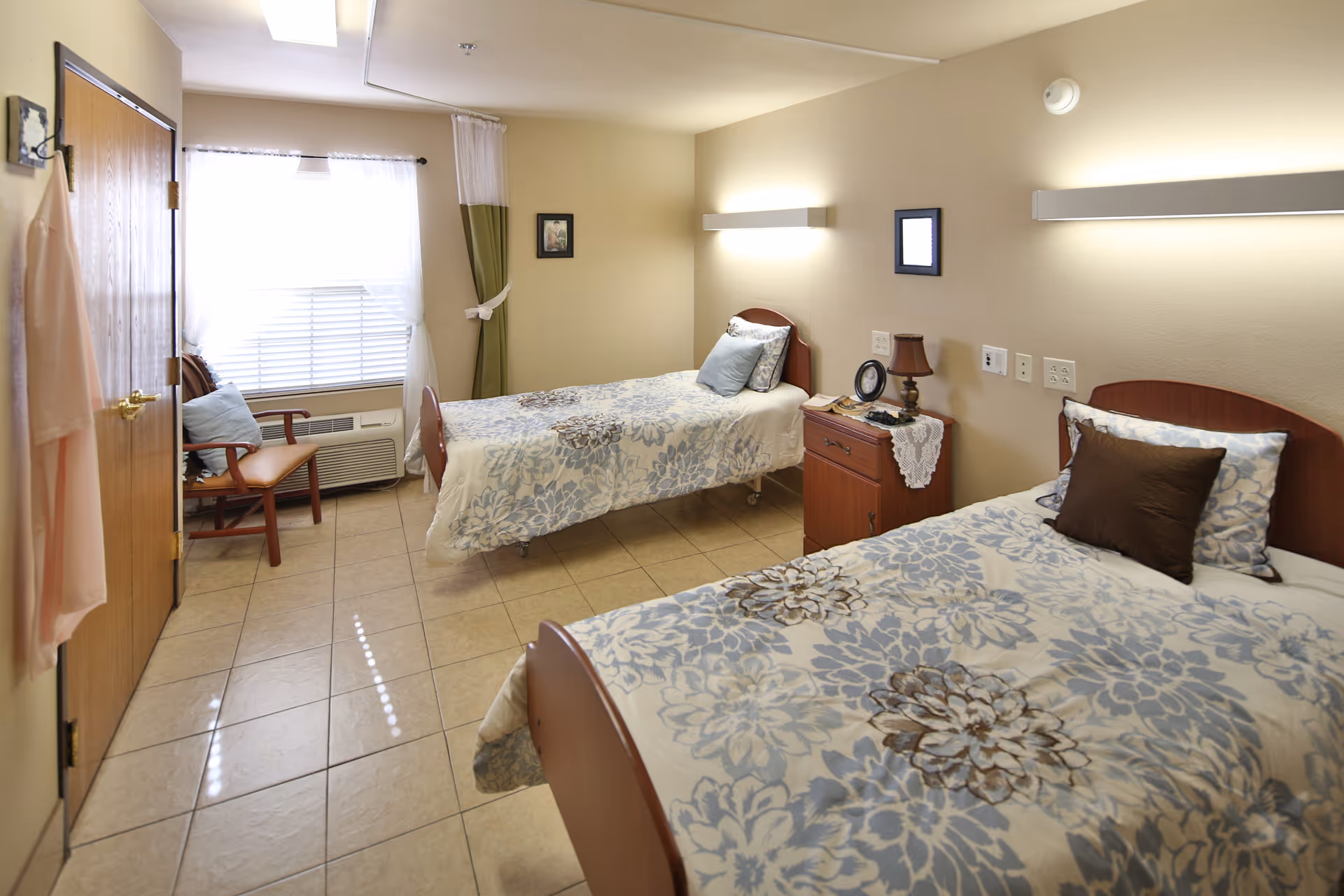 A clean and well-lit nursing facility bedroom with two single beds, each with floral patterned bedding and pillows. There is a wooden nightstand between the beds with a lamp and a small clock. A wooden chair with a cushion is placed near a window with white and green curtains. The room has tiled flooring and beige walls with two wall-mounted lights above the beds.