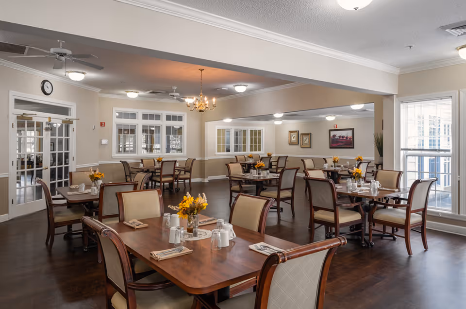 A well-lit dining room with multiple wooden tables and cushioned chairs arranged neatly. Each table has a small floral centerpiece with yellow flowers, napkins, and glassware. The room features large windows allowing natural light, ceiling fans, chandeliers, and framed artwork on the walls. The flooring is dark wood, and there are white double doors on one side.