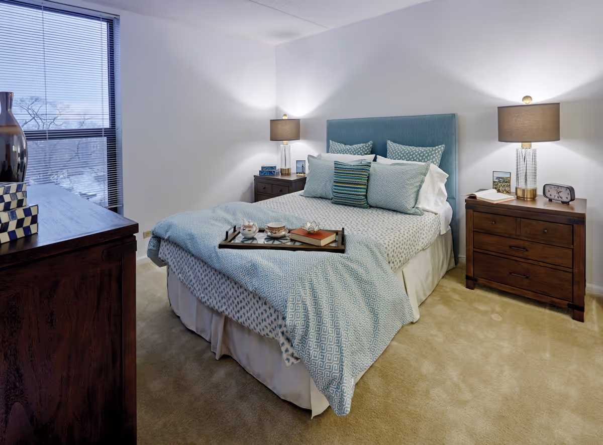 A cozy bedroom with a neatly made bed featuring blue and white patterned bedding and multiple pillows. There are two wooden nightstands on either side of the bed, each with a lamp and decorative items. A window with blinds is on the left side, and a wooden dresser with decorative objects is partially visible in the foreground.