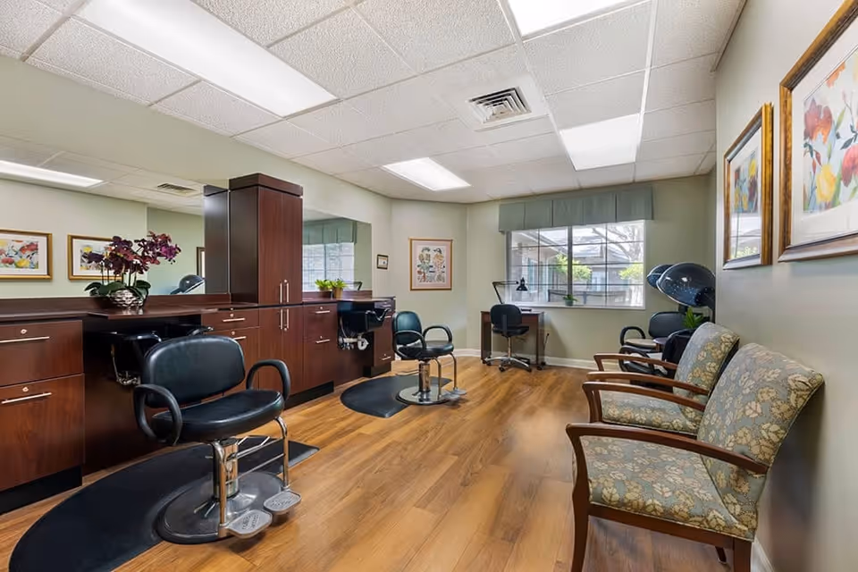 Interior view of a salon area in a senior living facility with wooden flooring, two black salon chairs in front of a large mirror and wooden cabinetry, a desk with a chair near a window, two floral upholstered chairs along the right wall, and hair drying stations in the background.