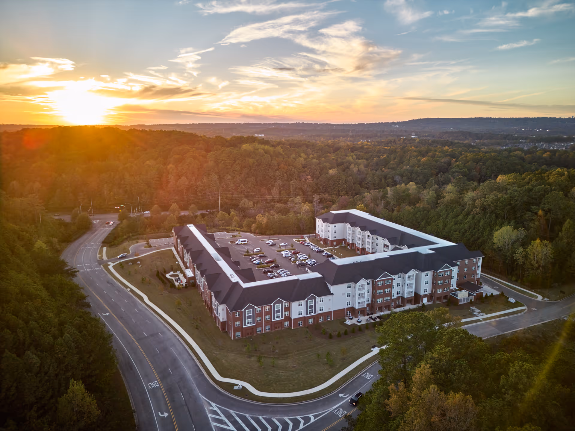 Aerial view of The Crossings at Riverchase senior living facility at sunset, showing a large U-shaped building surrounded by trees and parking lots, with a road curving around the property.