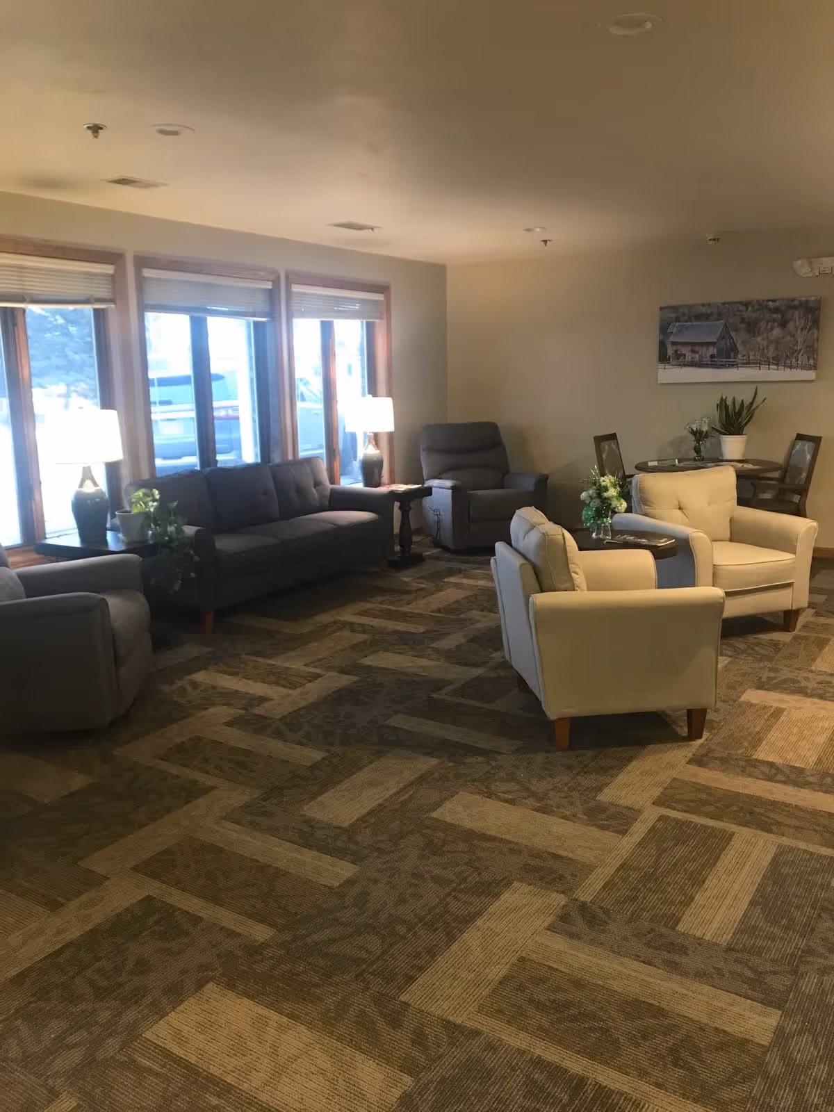 A cozy living room area with multiple seating options including a dark gray sofa, two light beige armchairs, and a gray recliner. There are two side tables with lamps and plants, large windows letting in natural light, a patterned carpet, and a round dining table with chairs and a plant on it. A painting of a barn hangs on the wall.