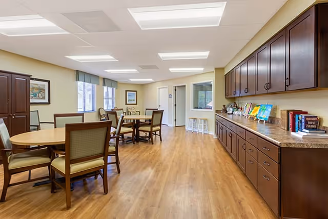 A bright and spacious dining room with wooden floors, several round tables surrounded by chairs, large windows with green valances, and a long countertop with dark wooden cabinets above and below. The walls are light-colored and decorated with framed artwork.