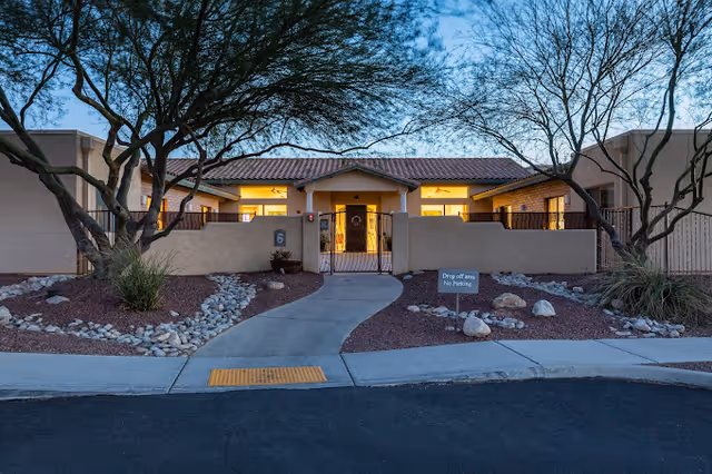 Exterior view of a single-story building at dusk with warm lights glowing from inside. The building has a tiled roof and beige stucco walls. A paved walkway leads to a gated entrance with a sign indicating 'Drop off area No Parking'. Desert landscaping with rocks and trees surrounds the walkway.