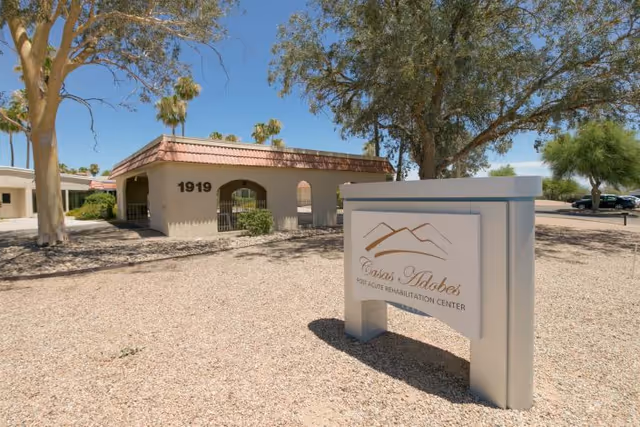 Exterior view of Casas Adobes Post Acute Rehabilitation Center showing a single-story building with the number 1919 on the wall, surrounded by trees and desert landscaping with gravel ground. A sign with the facility's name and logo is prominently displayed in the foreground.