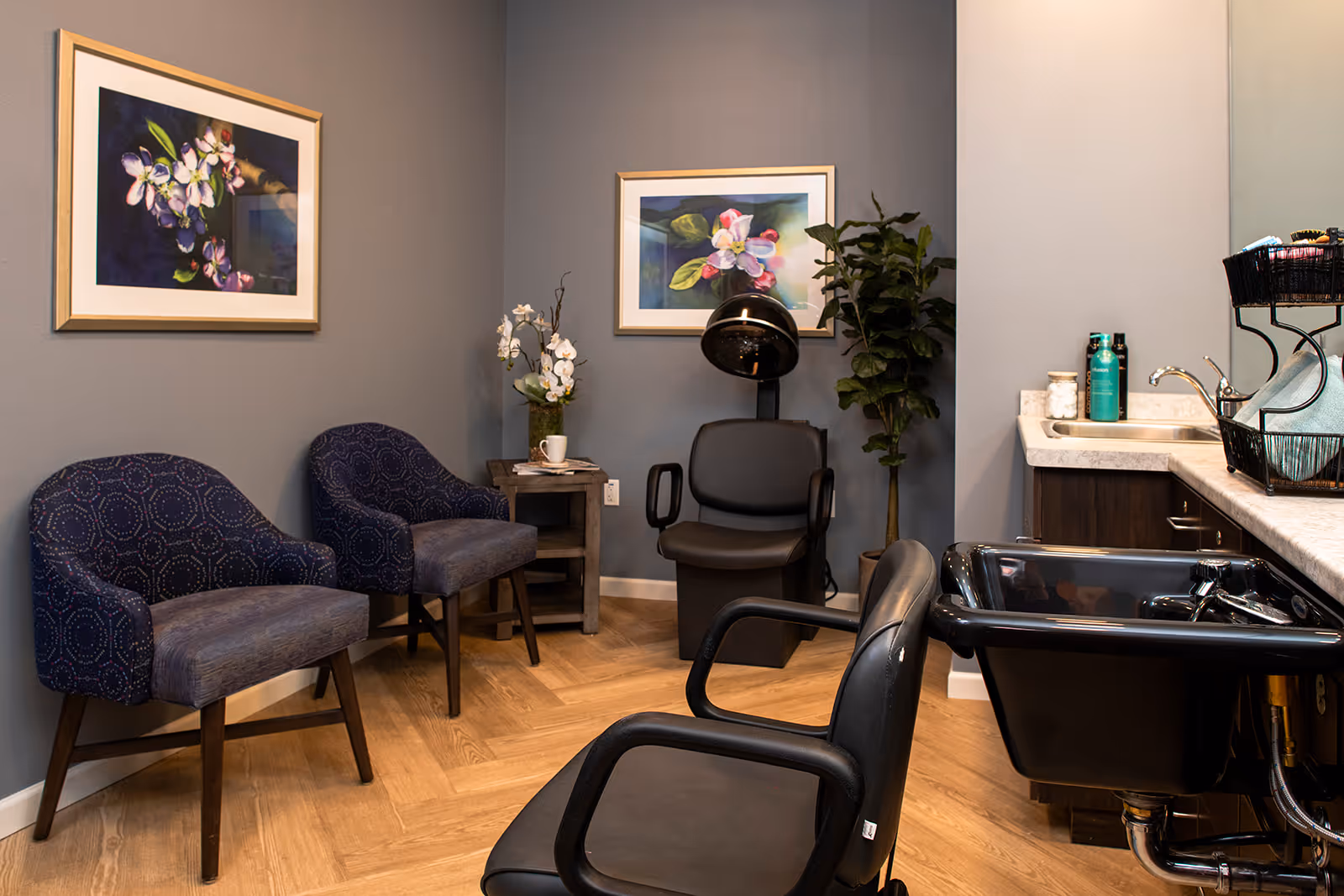 Interior of a hair salon area in a senior living facility with two blue patterned chairs, a black salon chair, a black hair washing sink, a hair dryer chair, a small side table with a flower vase and cup, two framed floral paintings on the wall, and a countertop with hair care products and a sink.