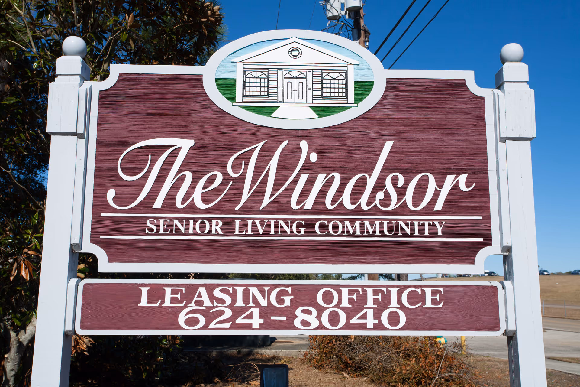 A large maroon wooden sign reading 'The Windsor Senior Living Community' with the leasing office phone number below.