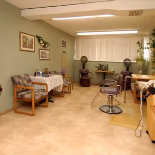 Interior room with salon chairs and hooded hair dryers, a small table with chairs, framed pictures and plants in a senior living community.