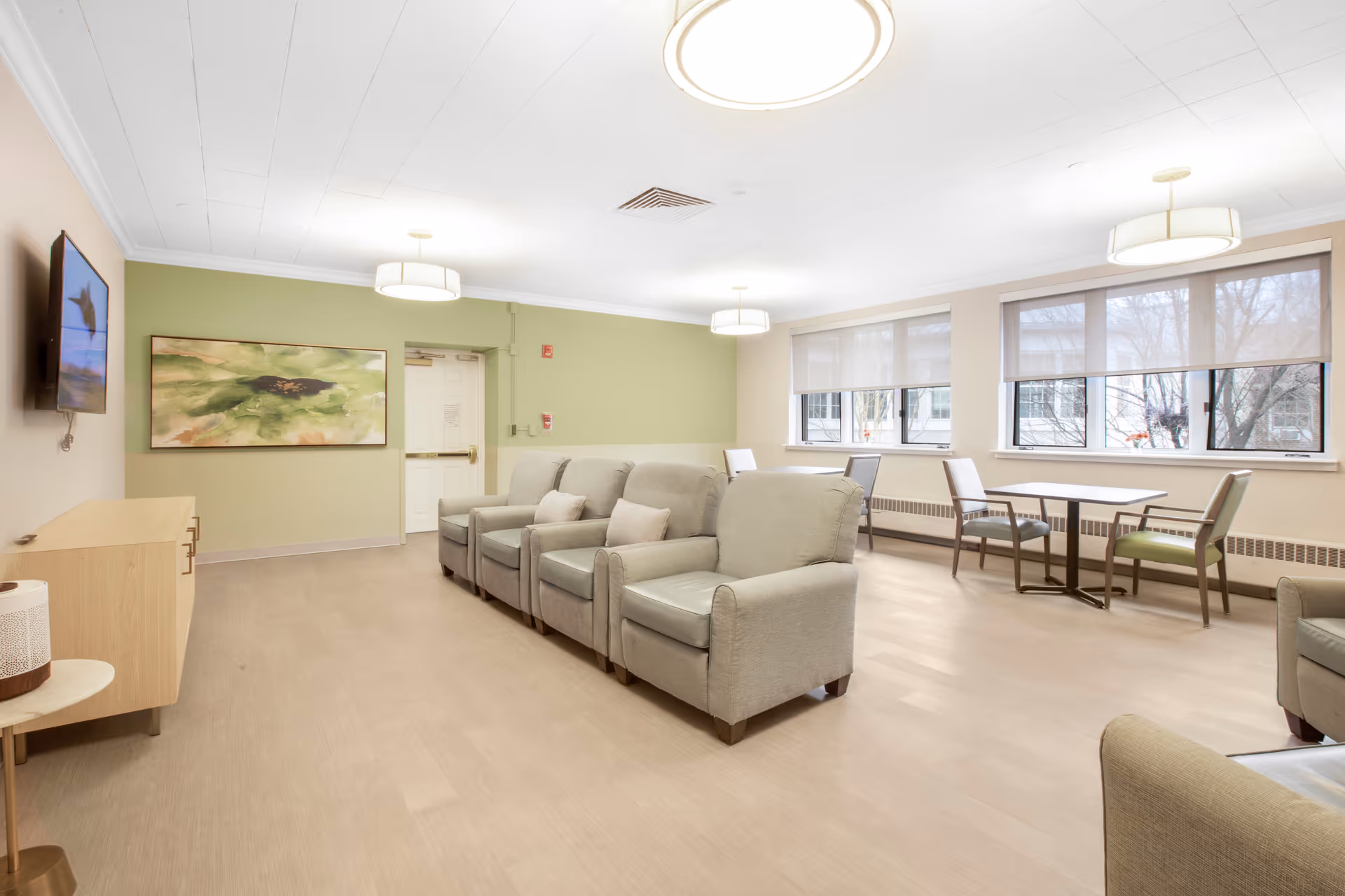 A bright and spacious senior living common area with beige armchairs arranged in a row facing a wall-mounted TV. The room has large windows with roller shades, light green and beige walls, modern ceiling lights, a wooden cabinet, and a small table with chairs near the windows.