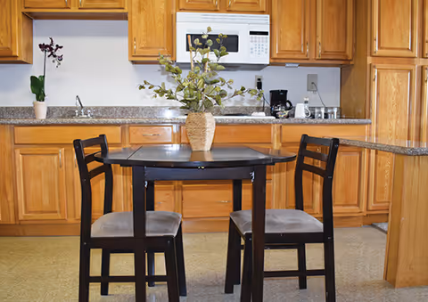 A small kitchen area with wooden cabinets, a granite countertop, a white microwave, a coffee maker, and a small black dining table with two chairs. A vase with green leafy branches is placed on the table.