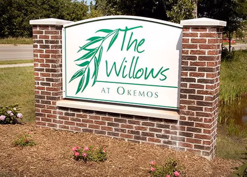A brick sign with white background displaying the text 'The Willows at Okemos' with a green willow branch graphic, situated outdoors with grass, flowers, and trees in the background.