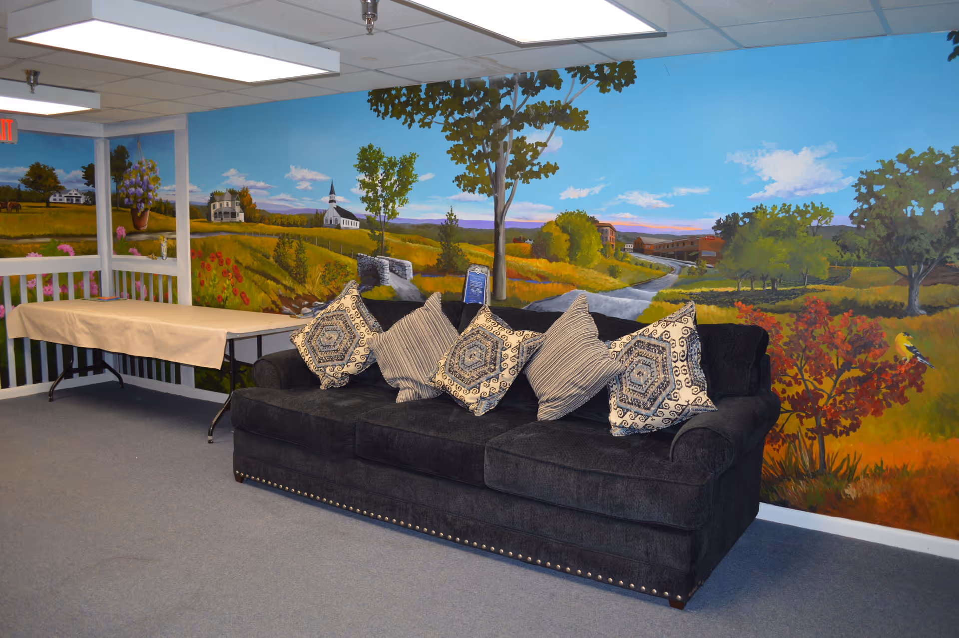 Dark upholstered couch with patterned throw pillows in a room with a large painted countryside mural and a covered folding table.
