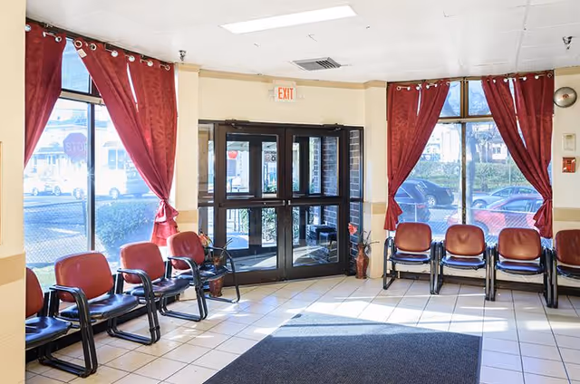 Sunlit entry lobby with double glass doors, red-cushioned chairs lining the walls, and red curtains at the windows.