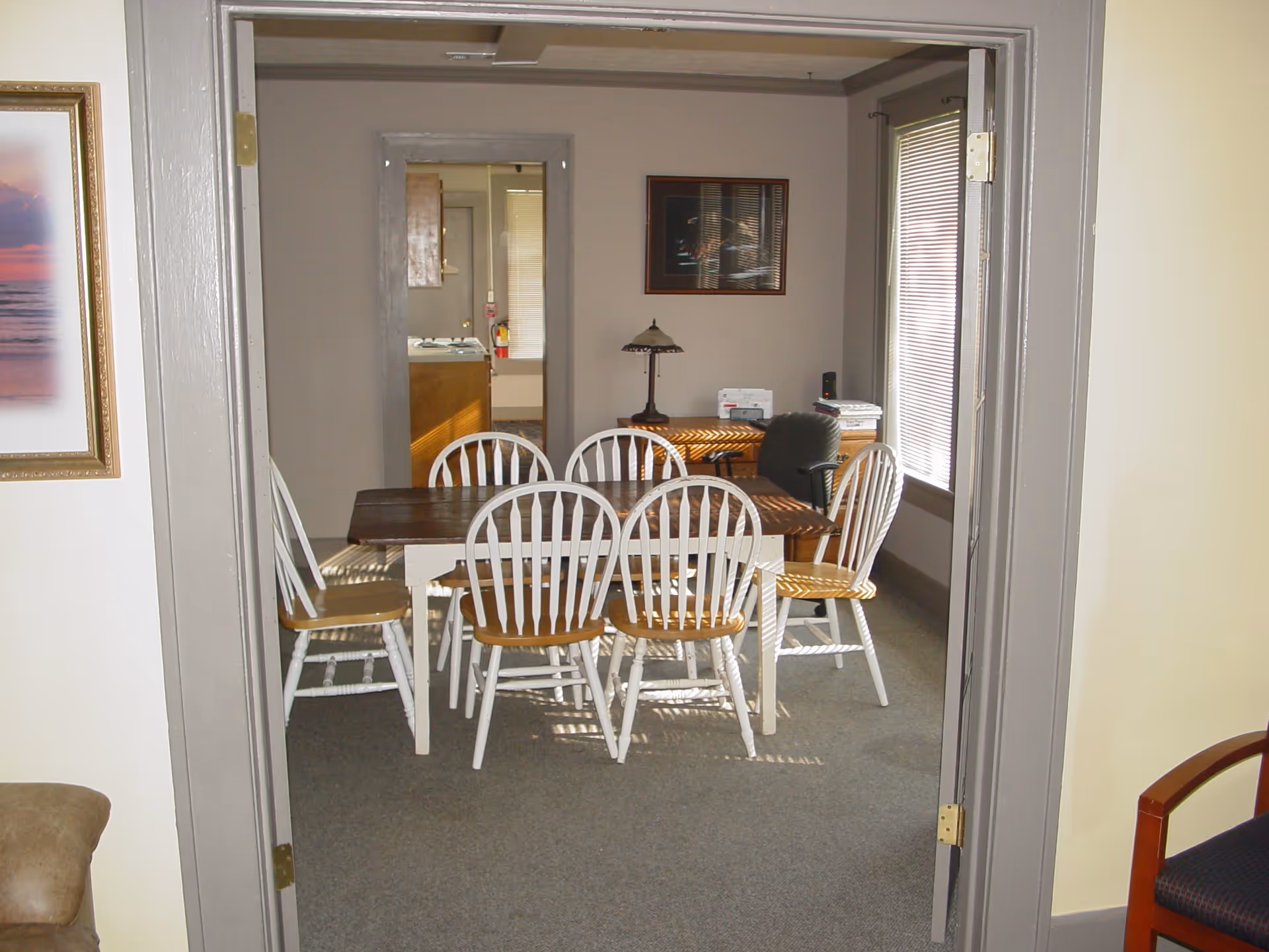 View through a doorway into a room with a wooden dining table surrounded by six white chairs with wooden seats. The room has gray walls and carpeted floor. A desk with a lamp, papers, and an office chair is against the far wall. There is a window with blinds on the right side and a framed picture on the wall.