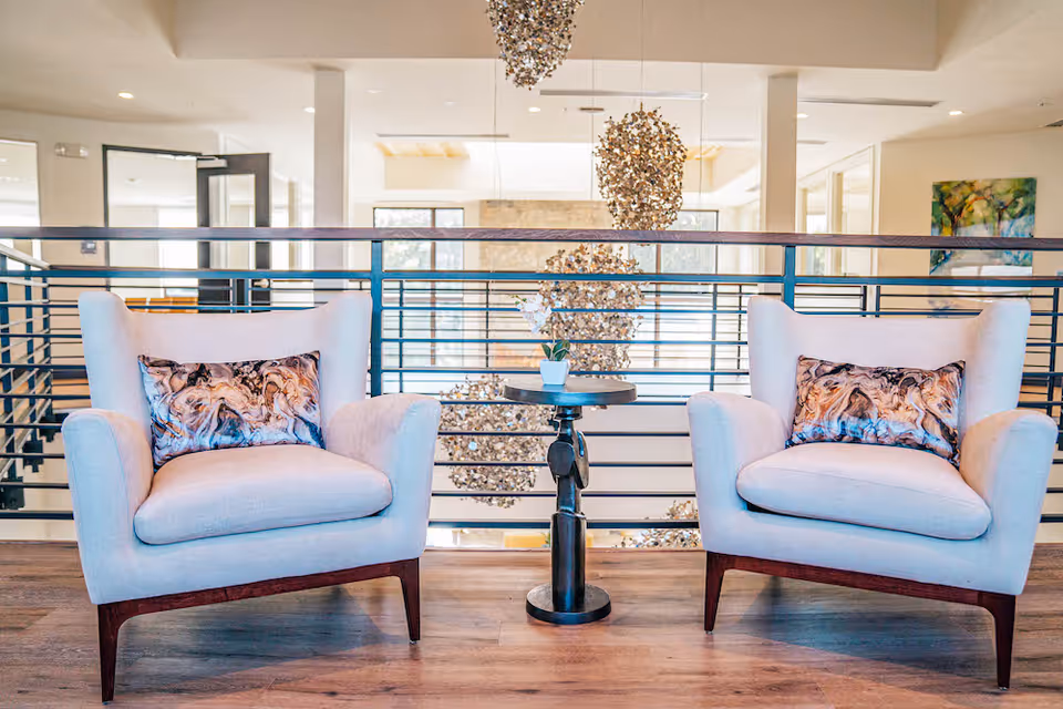 Two upholstered armchairs flanking a small round table on a wood floor with decorative hanging lights and a railing in the background.