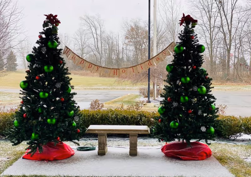 Two decorated Christmas trees flank a bench on a snowy outdoor patio with a 'Happy Holidays' banner and leafless trees beyond.