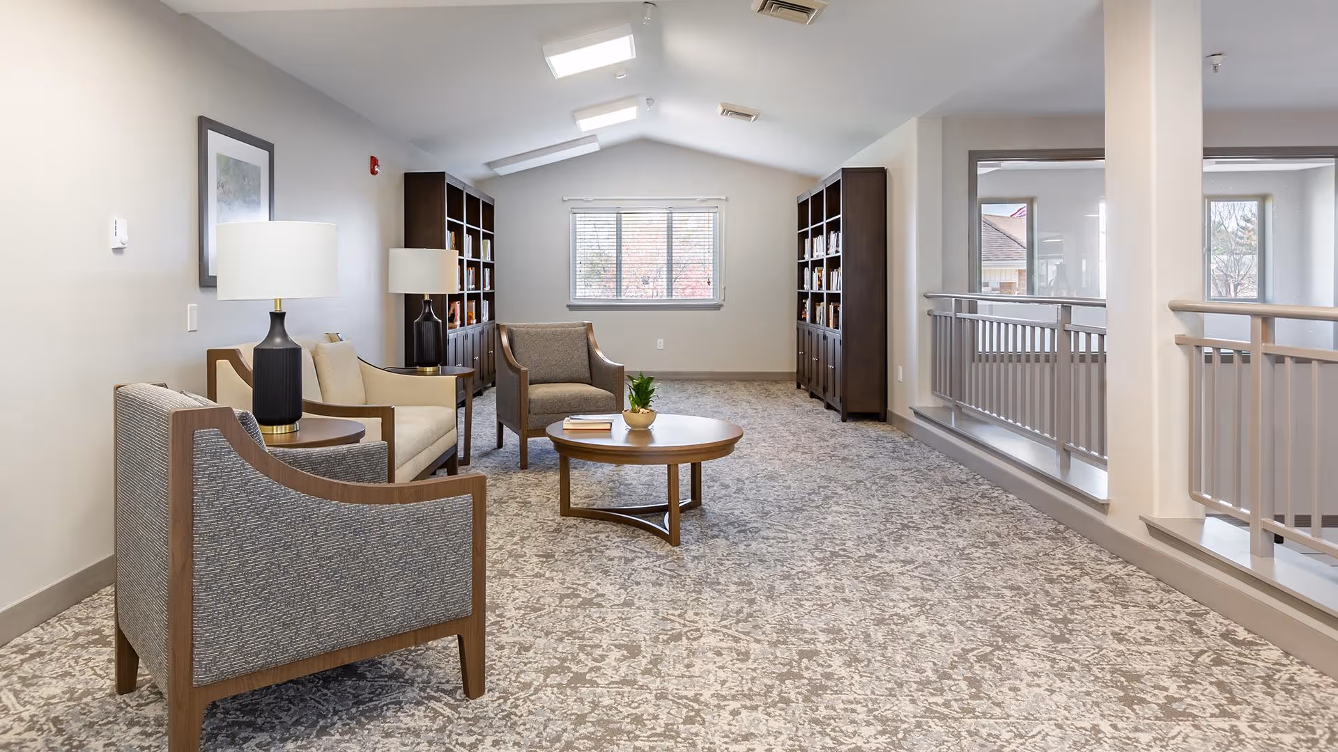 A bright and spacious sitting area with a patterned carpet, featuring two armchairs and a loveseat around a round wooden coffee table with a small plant and books. There are two tall dark wood bookshelves filled with books against the far wall, a window with blinds letting in natural light, and two table lamps on side tables. The right side has a railing overlooking another area below.