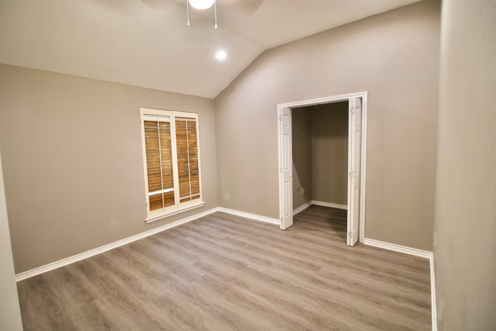 Empty room with light gray walls and wood-look flooring. There is a window with wooden blinds on the left wall and an open closet with white folding doors on the right wall. The ceiling is vaulted with a ceiling fan and light fixture.