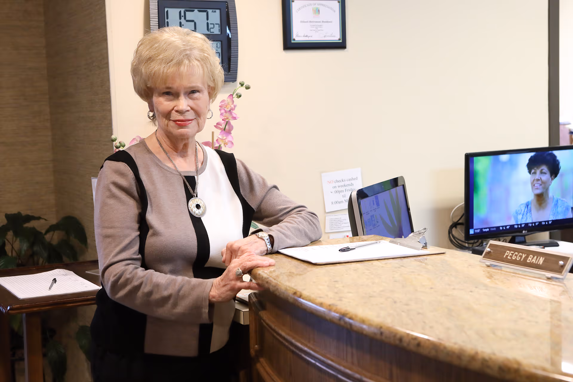 An elderly woman with short blonde hair stands behind a reception desk in a well-lit room. The desk has a nameplate that reads 'Peggy Bain,' a computer monitor displaying a woman on screen, a clipboard with papers, and a pen. Behind her on the wall is a digital clock showing 1:57 AM and a framed certificate. There are some plants and flowers in the background.