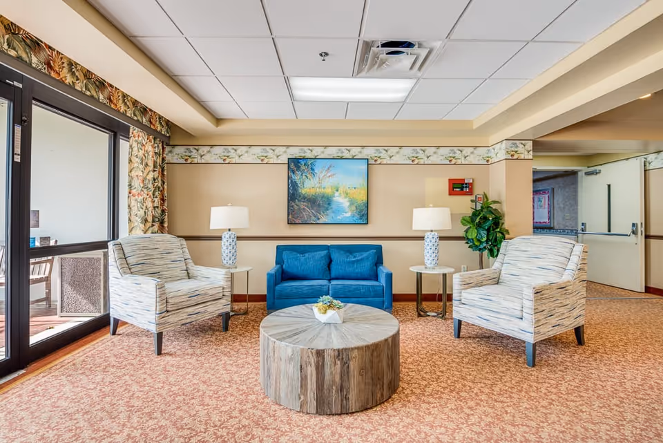 Bright lounge with a blue loveseat, two patterned armchairs, a round wooden coffee table and lamps beneath a framed painting.