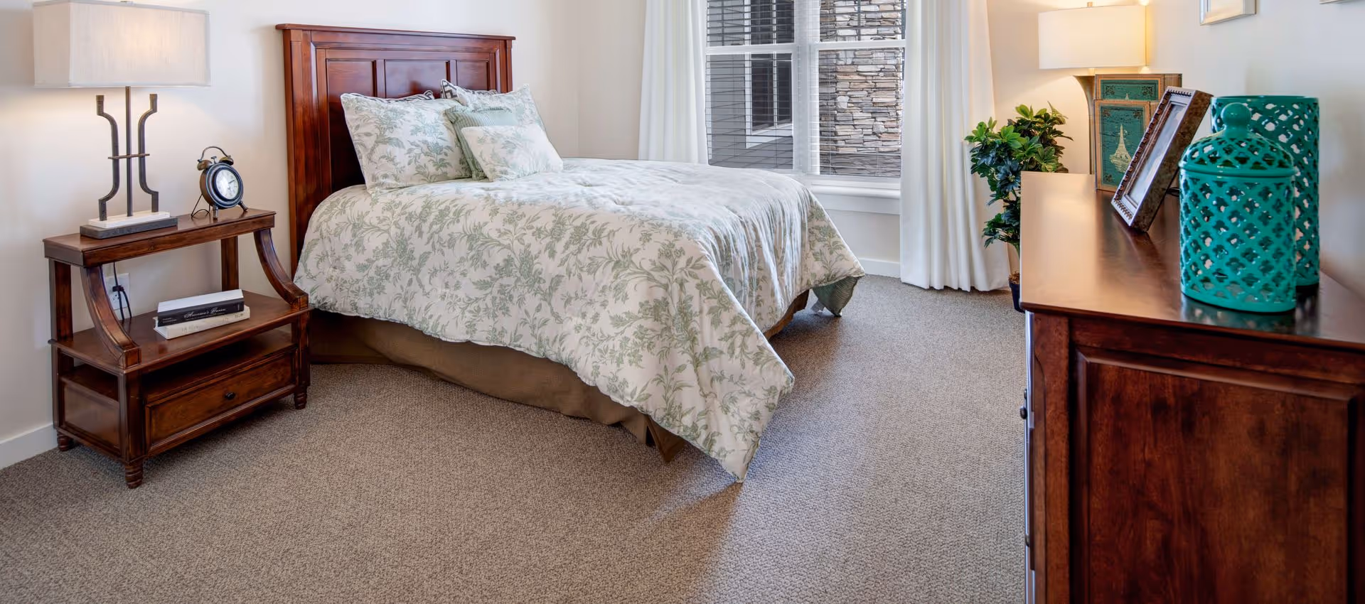 A cozy bedroom with a wooden bed frame and a bed dressed in white and green floral bedding. There is a wooden nightstand with a lamp and an alarm clock on the left side of the bed. On the right side, there is a wooden dresser with decorative items including a green ceramic jar, picture frames, and a lamp. A window with white curtains lets in natural light, and a potted plant is placed near the dresser.