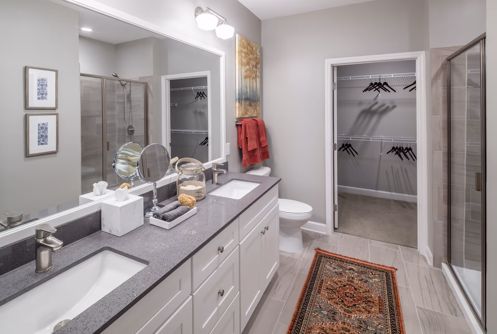 A modern bathroom featuring a double sink vanity with gray countertops, a large mirror, and two framed pictures on the wall. There is a glass-enclosed shower on the right side and a toilet next to a doorway leading to a walk-in closet with hangers. A decorative rug is placed on the tiled floor, and red towels hang on a rack above the toilet.
