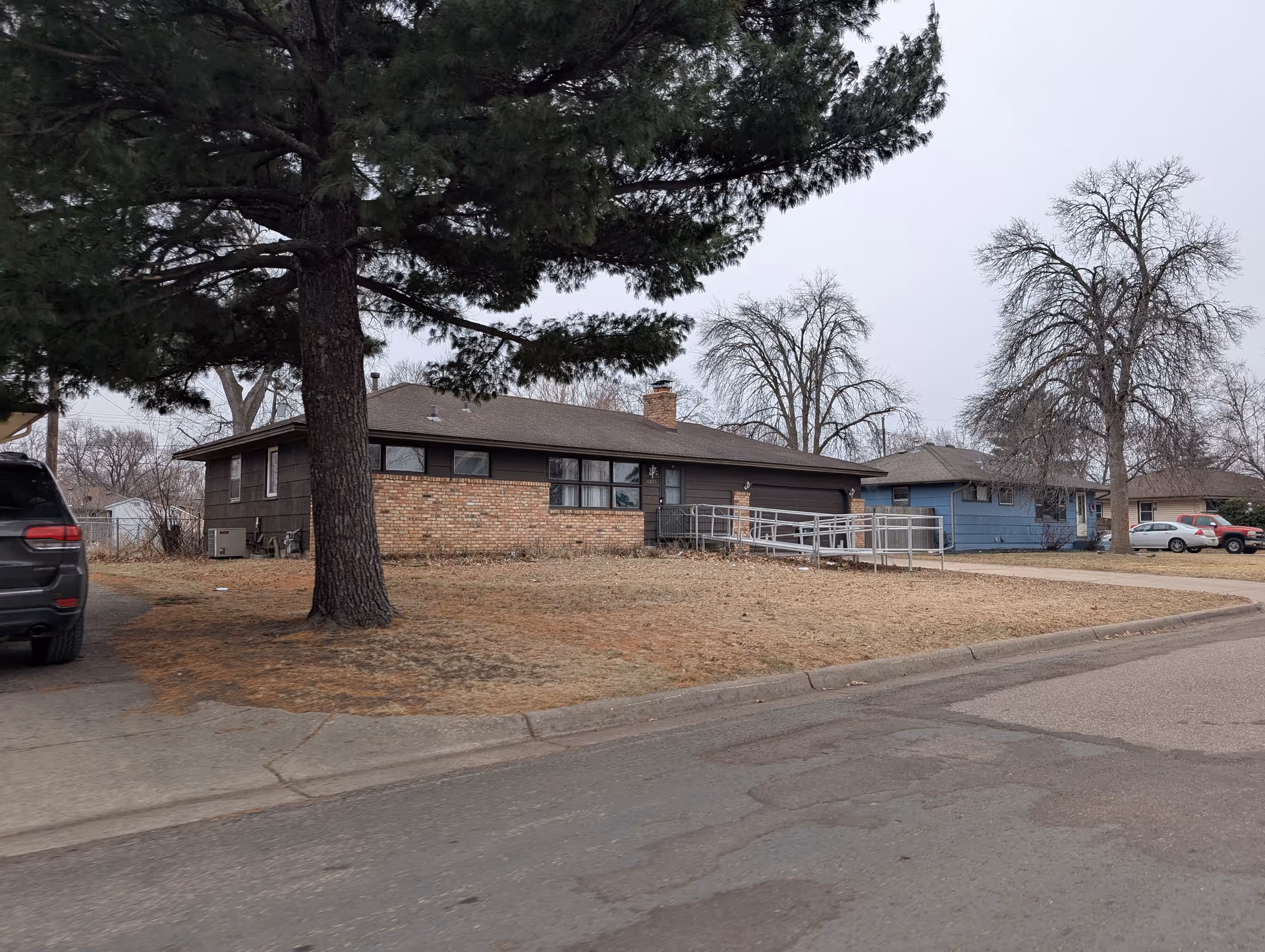 Exterior view of a single-story residential building with a brick and dark brown facade, featuring a wheelchair accessibility ramp leading to the front door. There are leafless trees and a large evergreen tree on the lawn, with a few parked cars visible on the street and driveway.