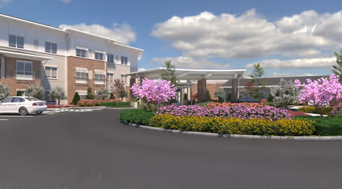 Exterior view of a senior living facility named Westhill Newnan Crossing featuring a circular driveway with a landscaped roundabout filled with colorful flowers and small trees. The building has a brick and light-colored facade with multiple windows and a covered entrance. The sky is partly cloudy.